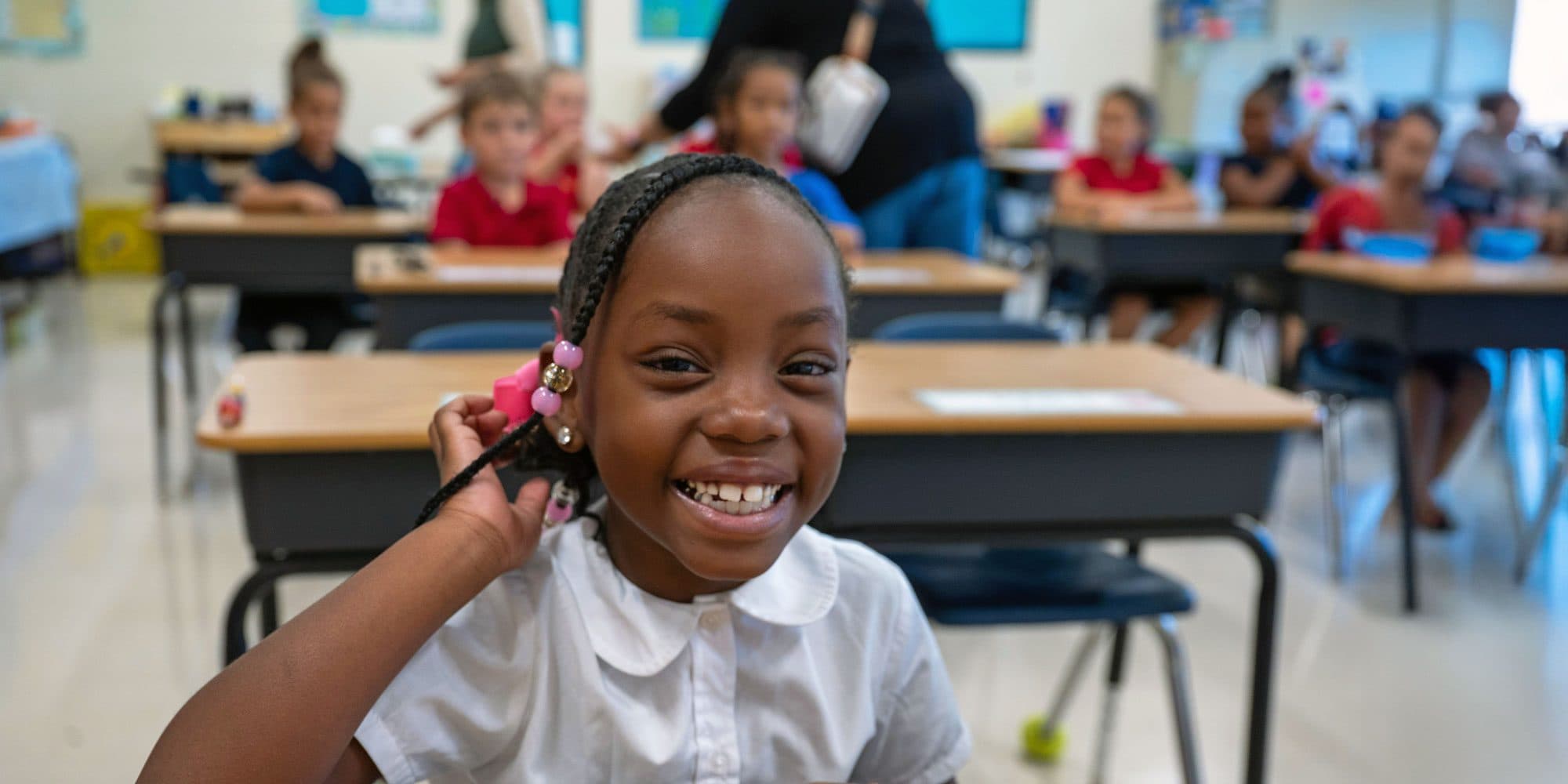 Smiling student at a classroom desk with classmates in the background.
