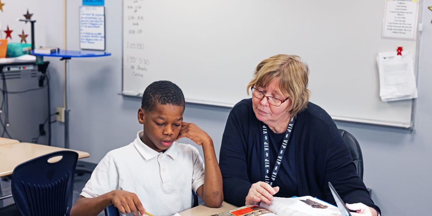 Student reading a book with an adult at a classroom table.